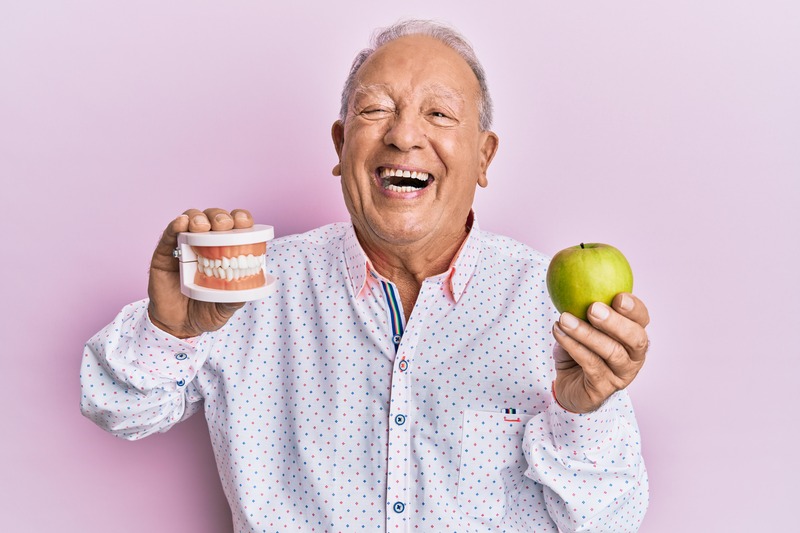 A laughing man holding a model denture and a green apple